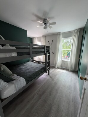 Una habitación con literas, ventilador de techo, ventana con cortinas y una puerta.