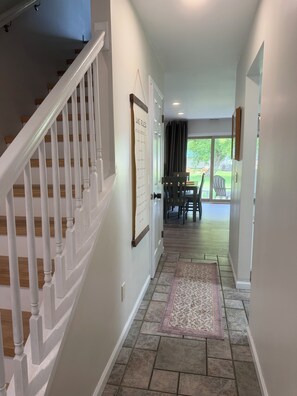 Un couloir avec un escalier en bois, un tapis et une porte donnant sur un espace repas avec des sièges à l’extérieur.