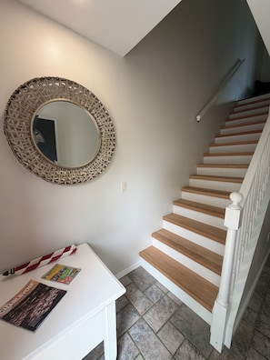 Un escalier avec des marches en bois et des rampes blanches, un miroir rond avec un cadre tressé, et une table blanche sur laquelle sont posés des magazines.