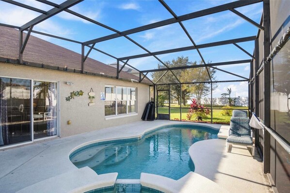 Una piscina exterior cubierta con cerramiento de vidrio, un patio y vistas a un jardín ajardinado.