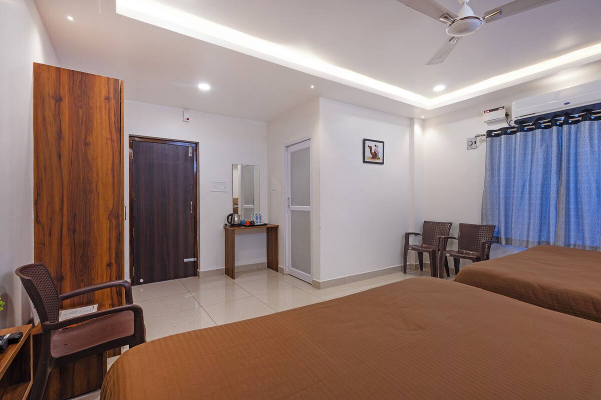 A hotel room with two beds, a desk with a chair, a wardrobe, a mirror, and a window with curtains.