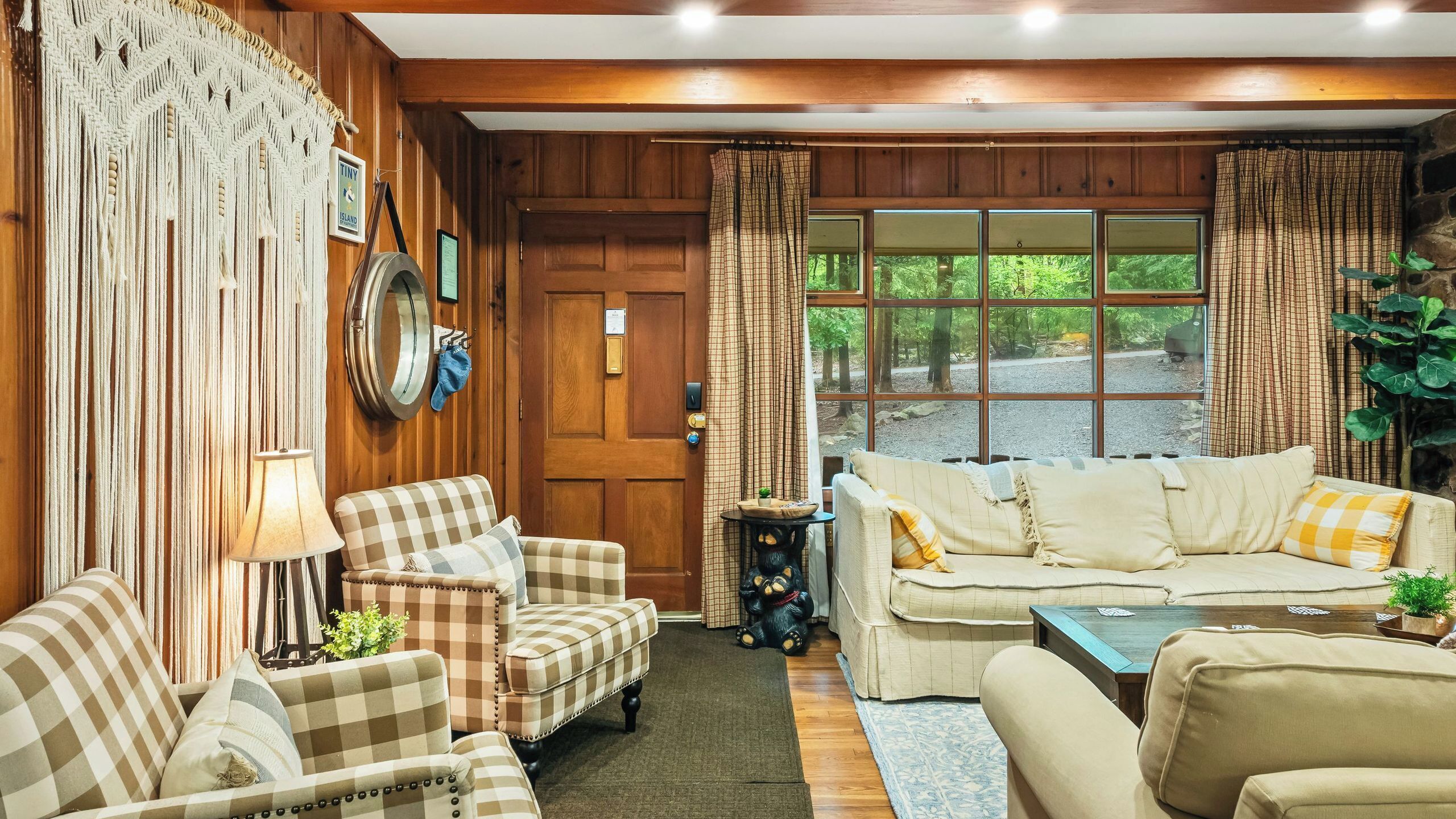 A cozy living room with a checkered sofa, armchairs, a wooden wall, and a large window with curtains.