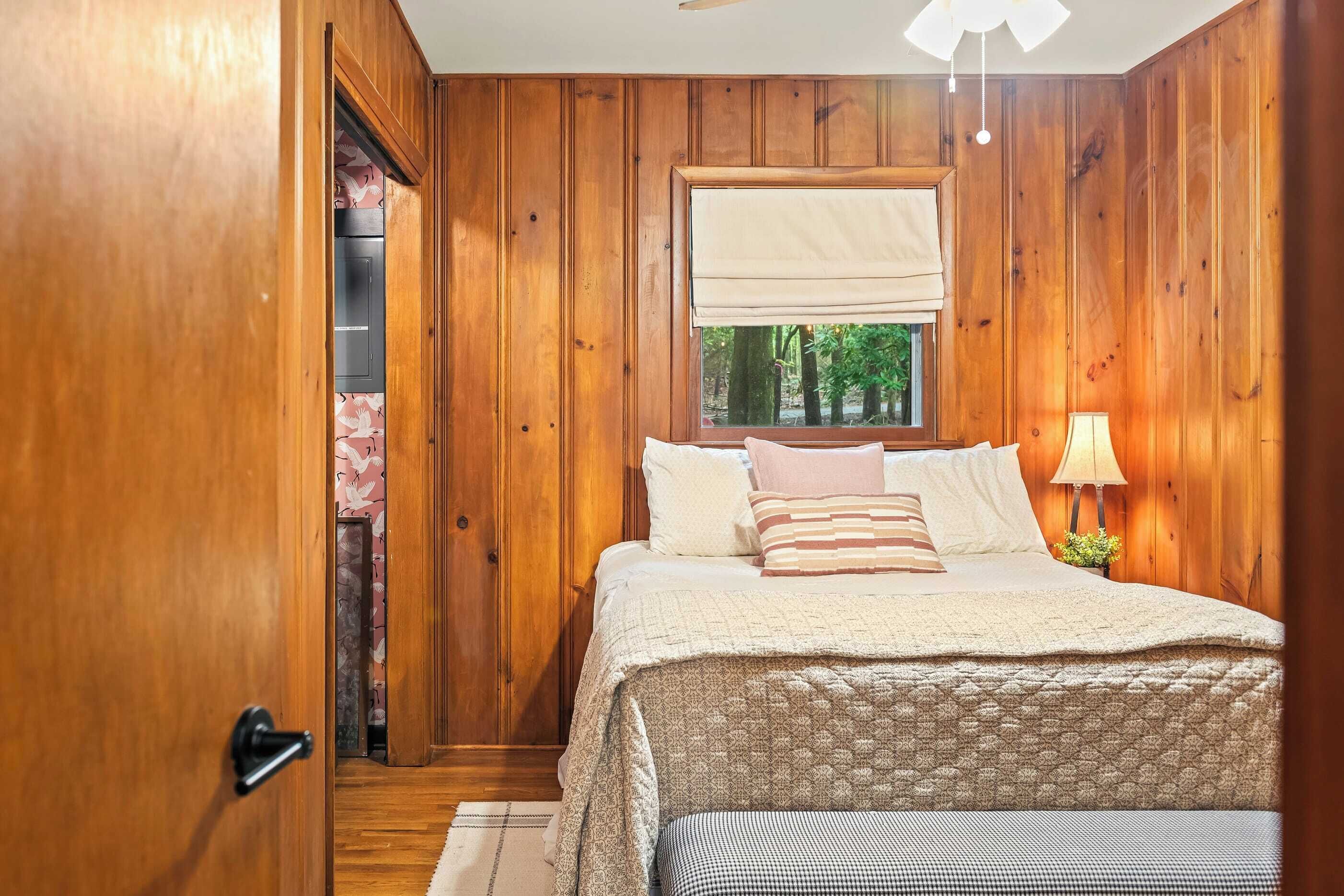 A wooden-paneled bedroom with a bed, a window with a view of trees, a lamp, and a door leading to another room.