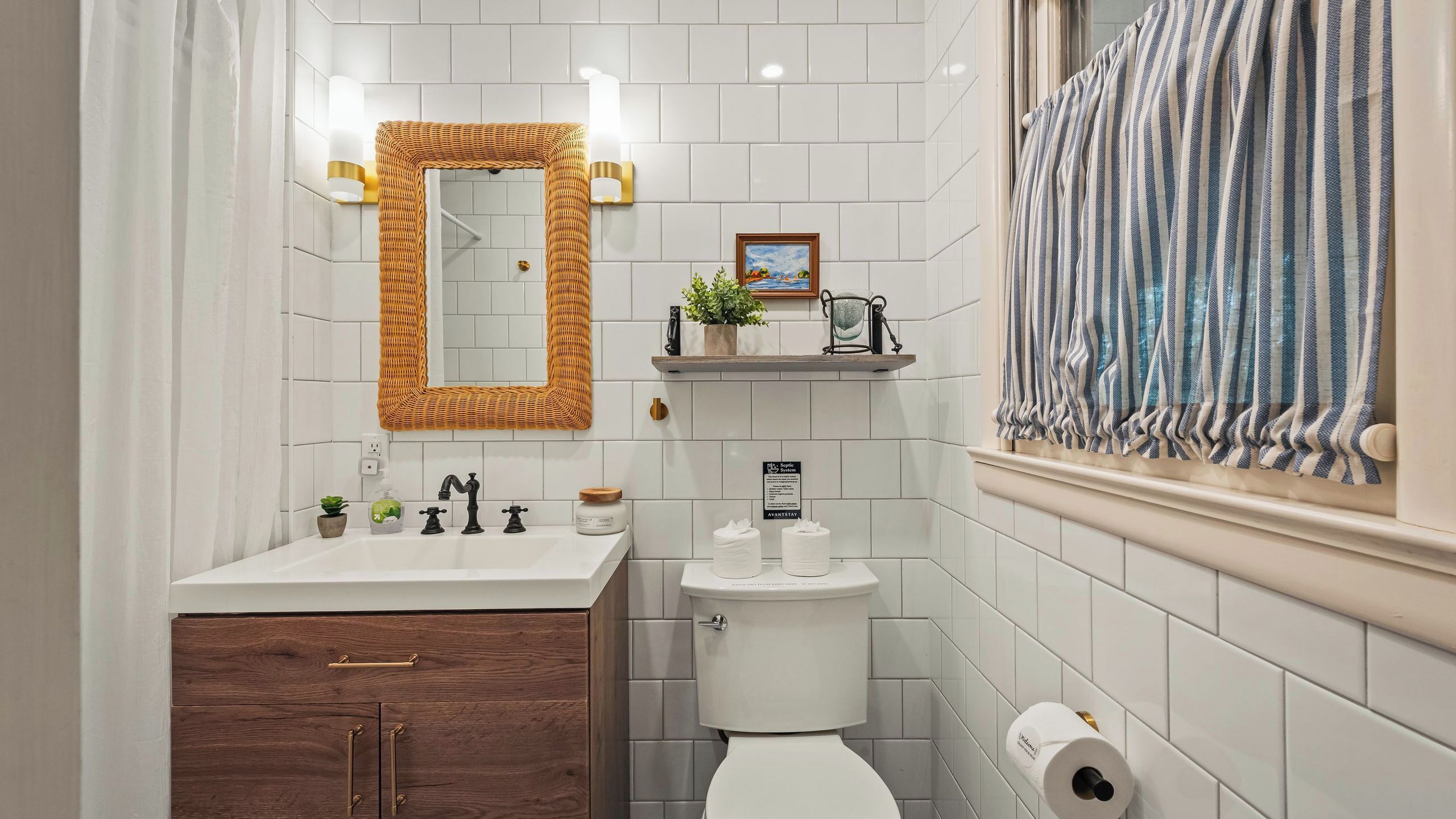 A bathroom with a white sink, a toilet, a mirror with a woven frame, a small shelf with a plant, and a window with striped curtains.