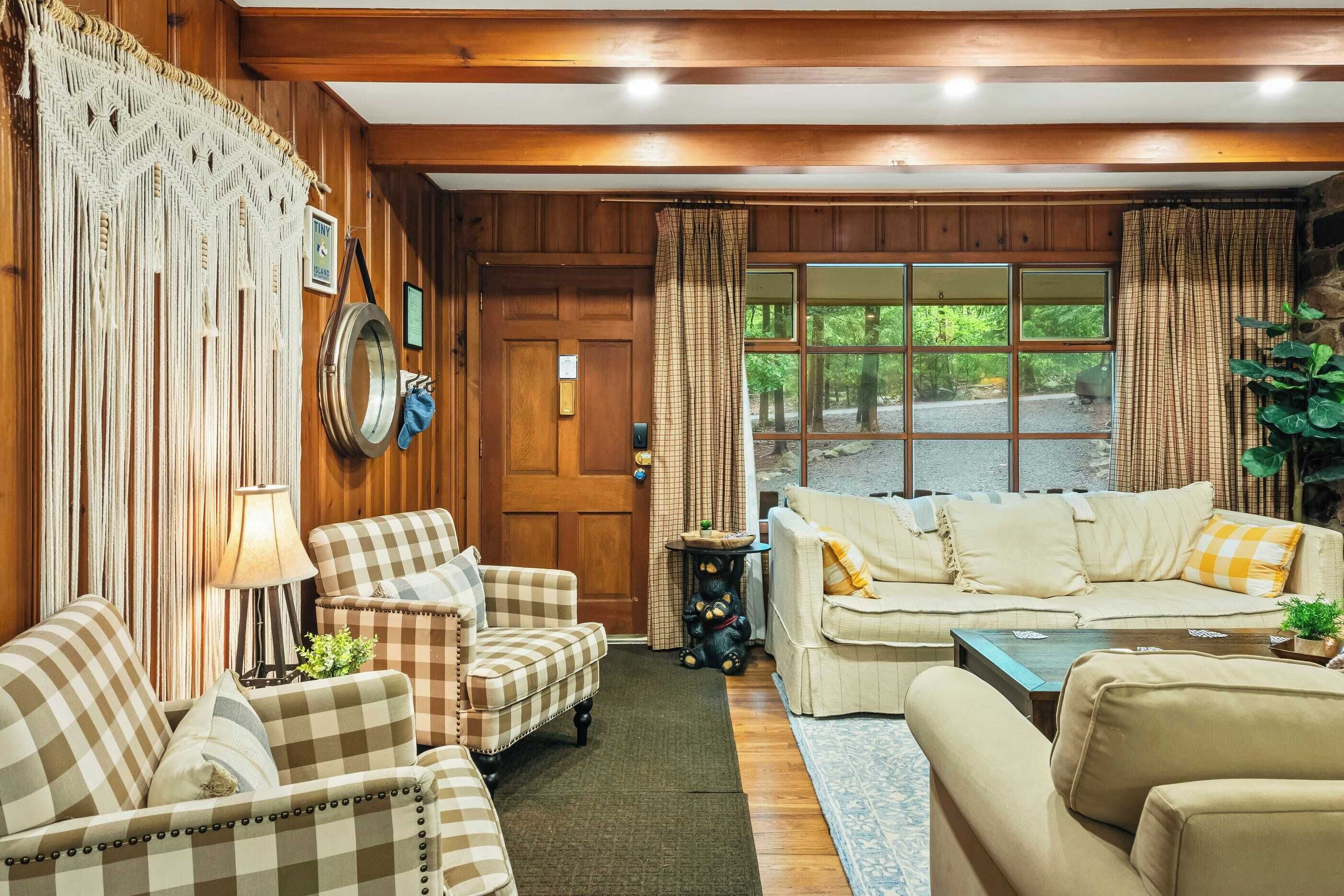 A cozy living room with wooden walls, checkered furniture, a large window with curtains, and a mirror on the wall.