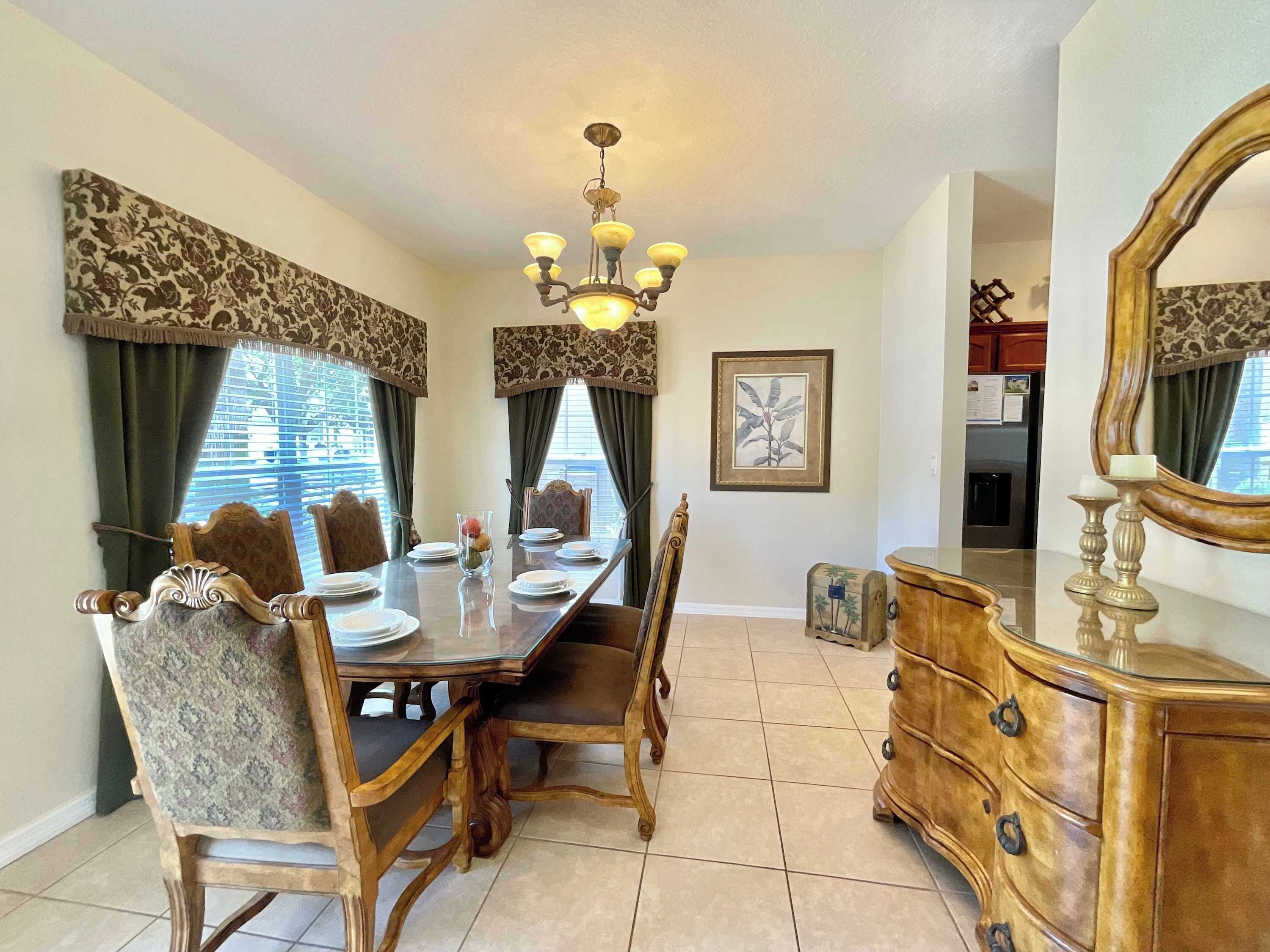 A dining area with a wooden table set for four, surrounded by ornate chairs. A chandelier hangs from the ceiling, and a framed botanical print is on the wall.