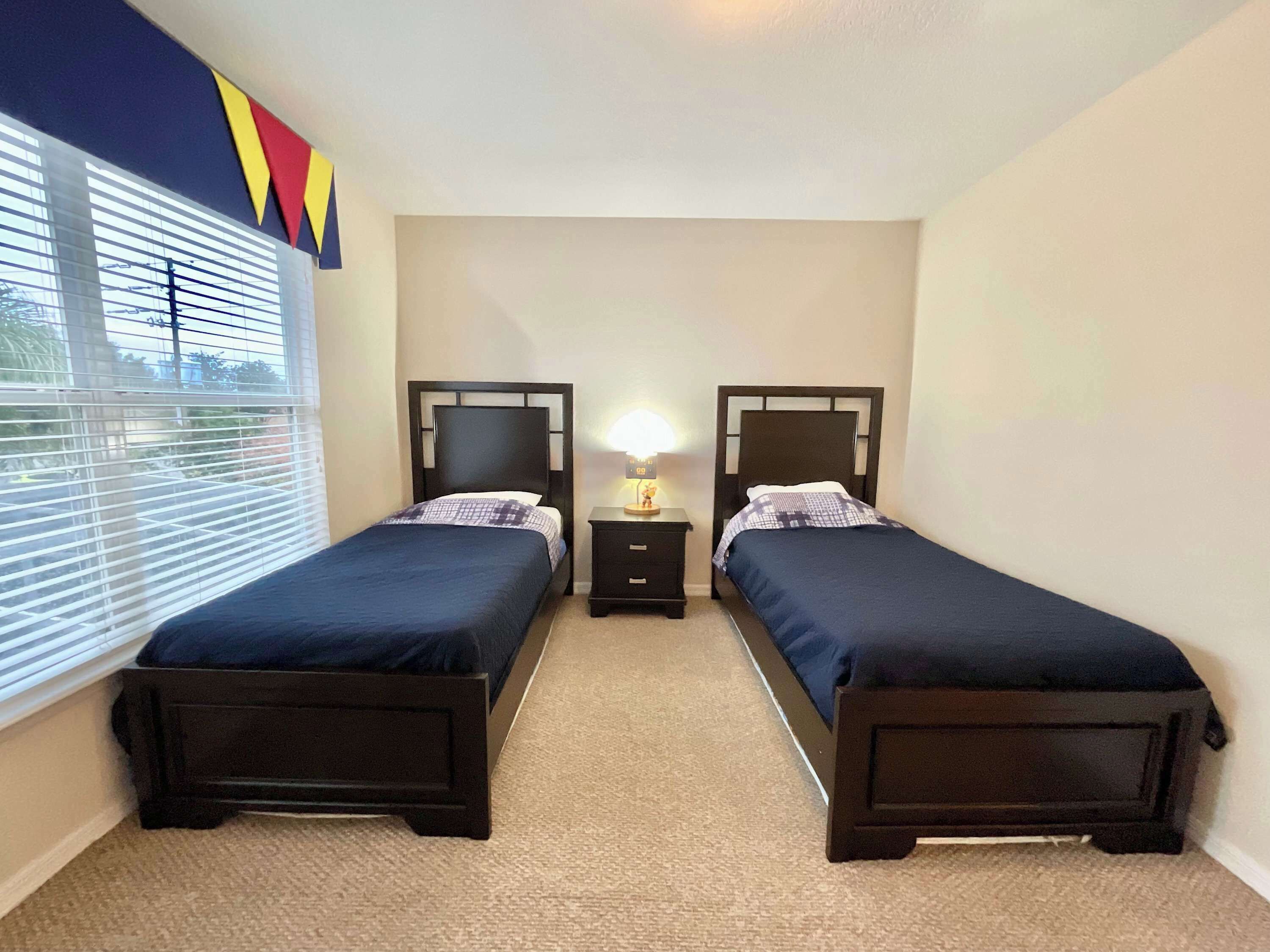 A bedroom with two single beds, a nightstand with a lamp, and a window with blinds.