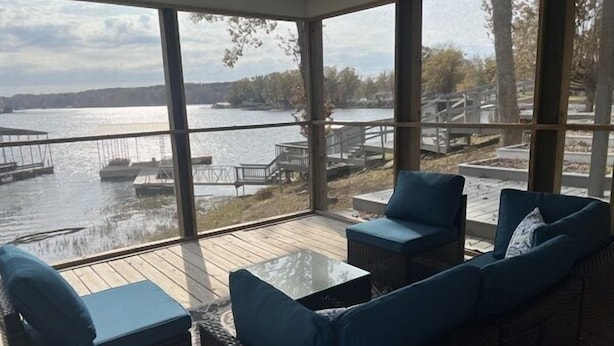 Uma sala de sol com grandes janelas com vista para um lago, mobiliada com sofás de vime e uma mesa de vidro.