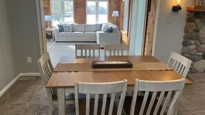 Uma sala de jantar com mesa de madeira e cadeiras brancas, um sofá ao fundo e uma parede de pedra com um abajur.