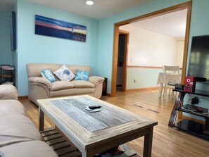 A living room with a beige sofa, a wooden coffee table, a TV stand with a flat-screen TV, and a painting on the wall. - Relaxing Harwich retreat with beautiful back yard/ pet friendly! (Harwich)