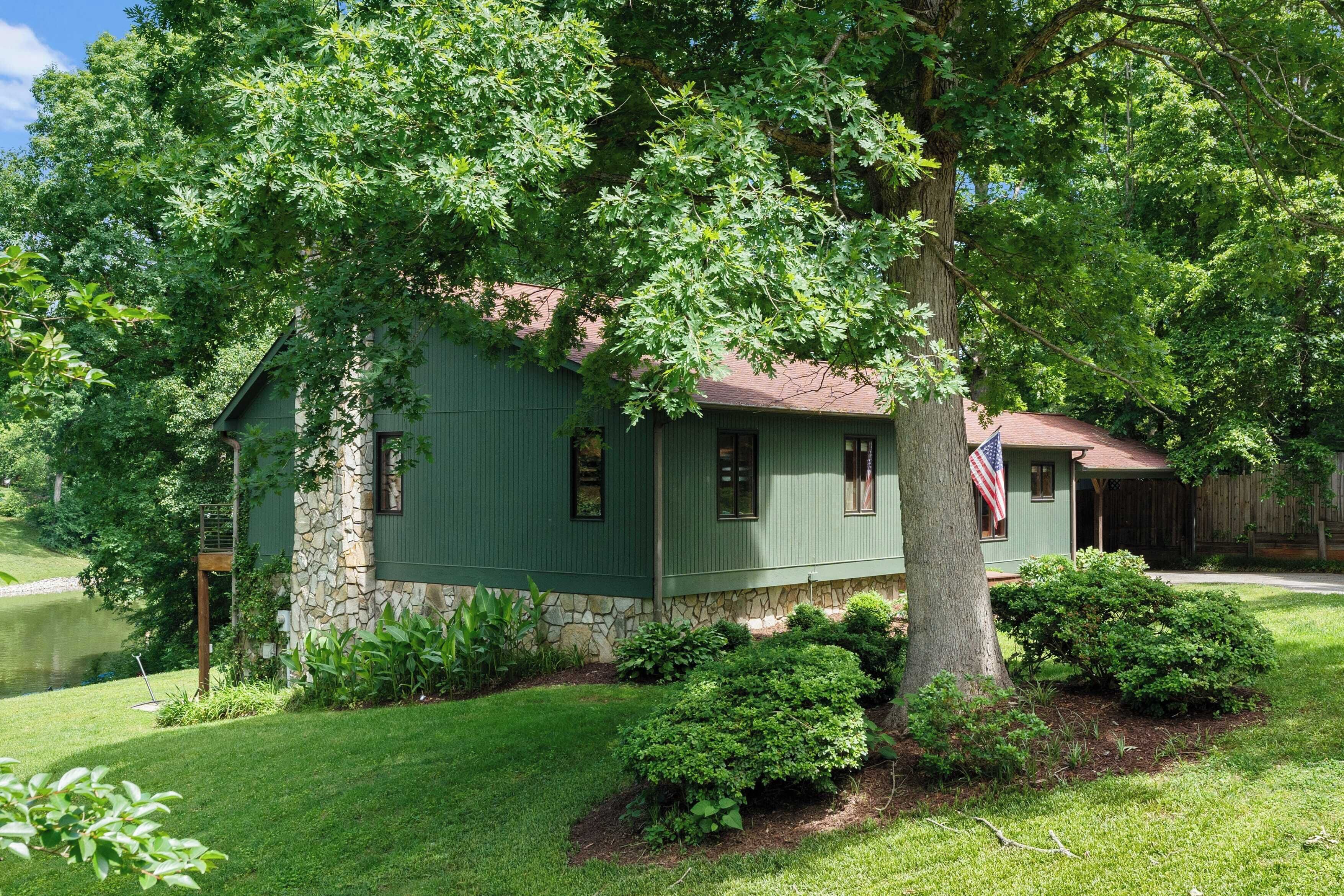 A green house with a stone foundation, surrounded by lush greenery and a small pond.