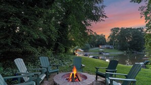 A circular fire pit with chairs around it, set in a garden with a view of a lake and a building in the background. - Shady Daze: Waterfront Home w/ Deck, Hot Tub & Lake Views (Sherrills Ford)