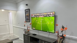 A living room with a flat-screen TV mounted on the wall, a dark wooden entertainment unit, and a vase with orange flowers. - 2-bedroom 2 Kings Bed. Breakfast in amazing Kasoa Gh with WiFi, AC. Sleep 2 (Kasoa Gh)