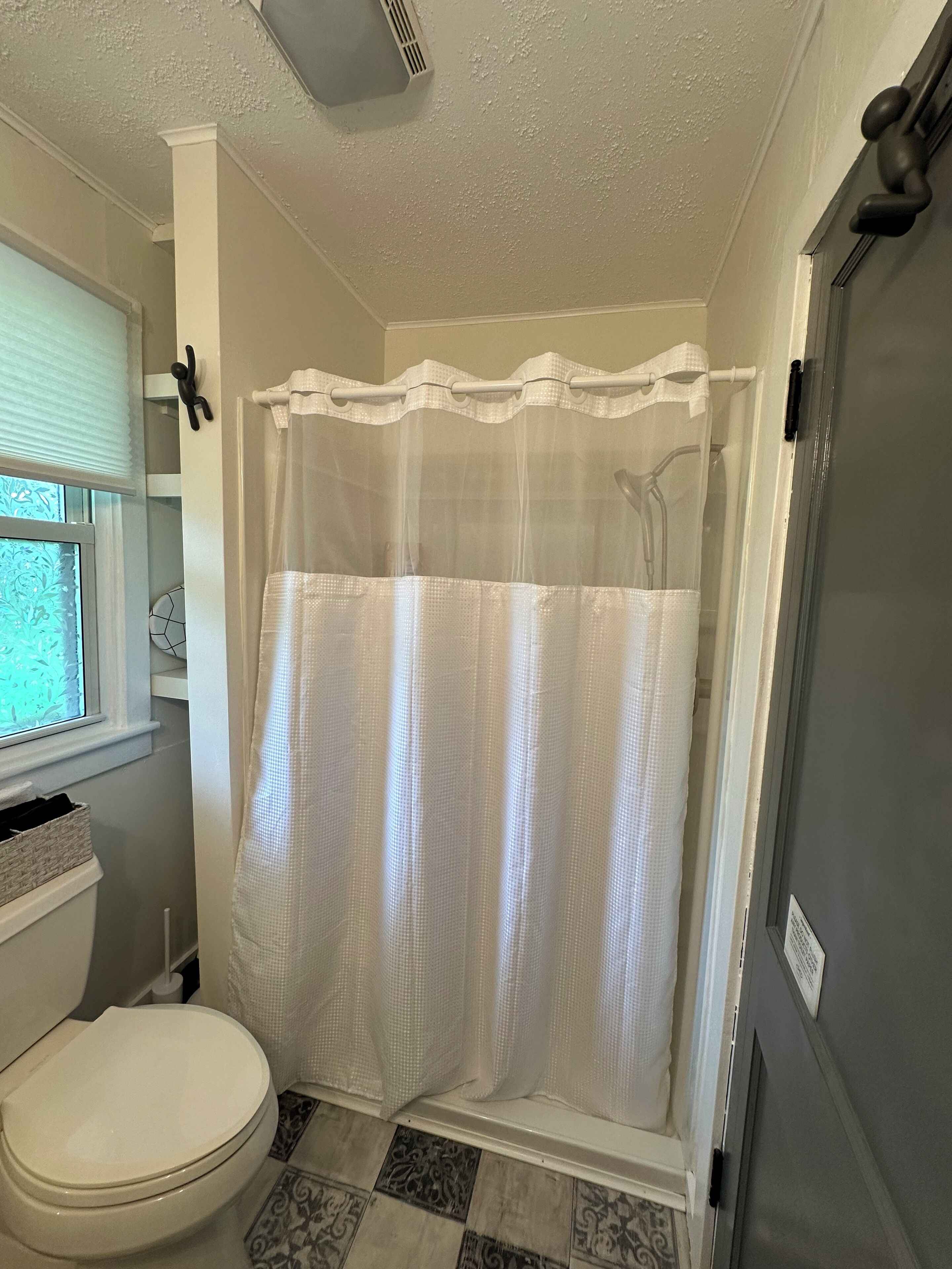 A bathroom with a toilet, a shower with a white curtain, and a window with blinds.