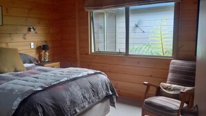 Een slaapkamer in een houten blokhut met een bed, een stoel, een nachtkastje, een lamp en een raam met uitzicht op bomen.