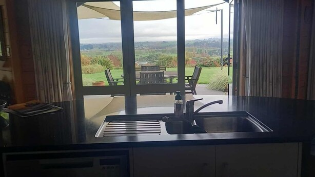 Een keuken met een spoelbak, een schaal fruit en uitzicht op een tuin en velden.