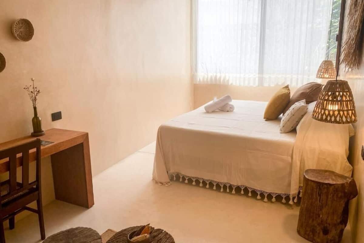 A bedroom with a bed, bedside table, lamp, and a window with sheer curtains.
