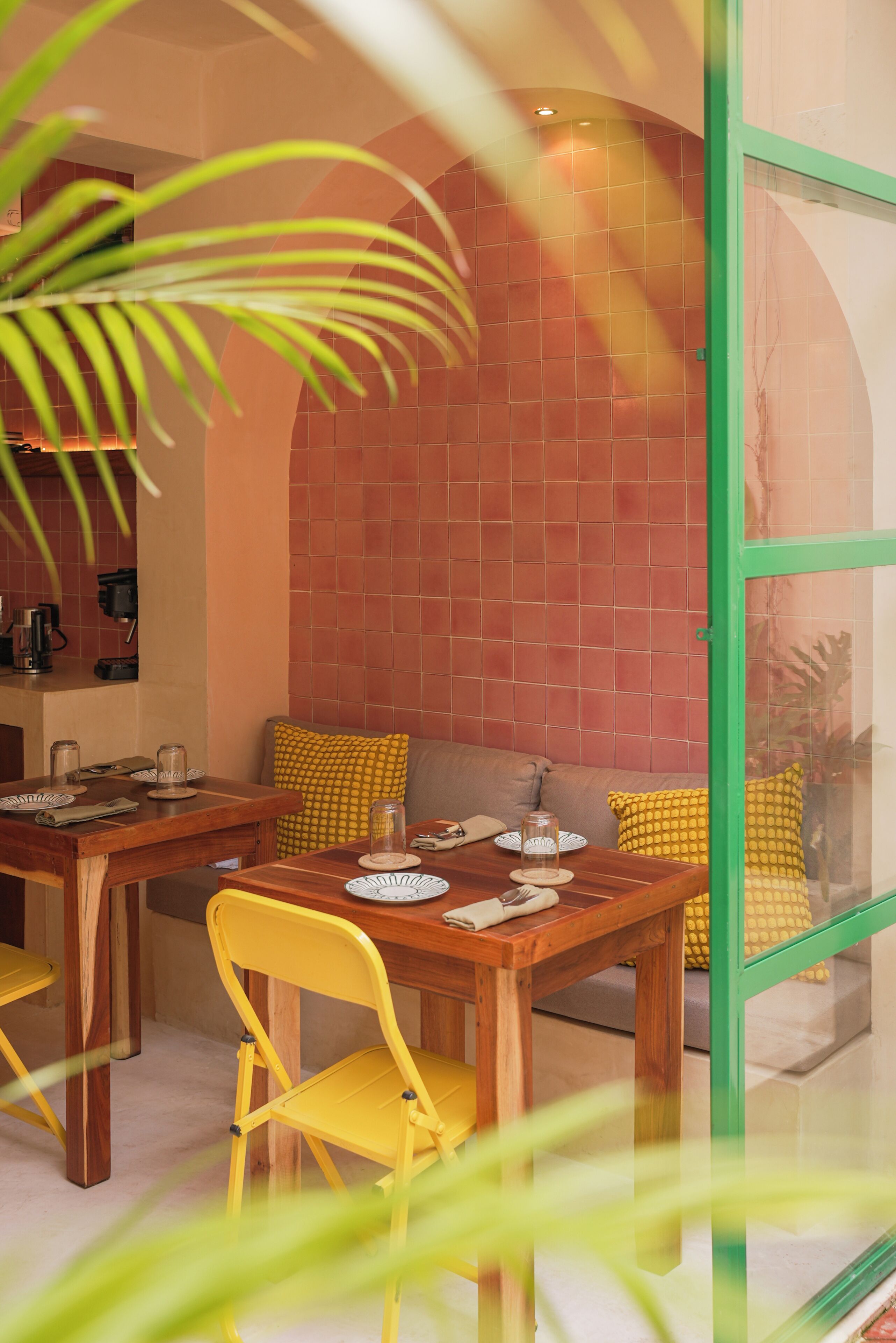 A dining area with a wooden table and yellow chairs, a red tiled wall, and a green-framed window.