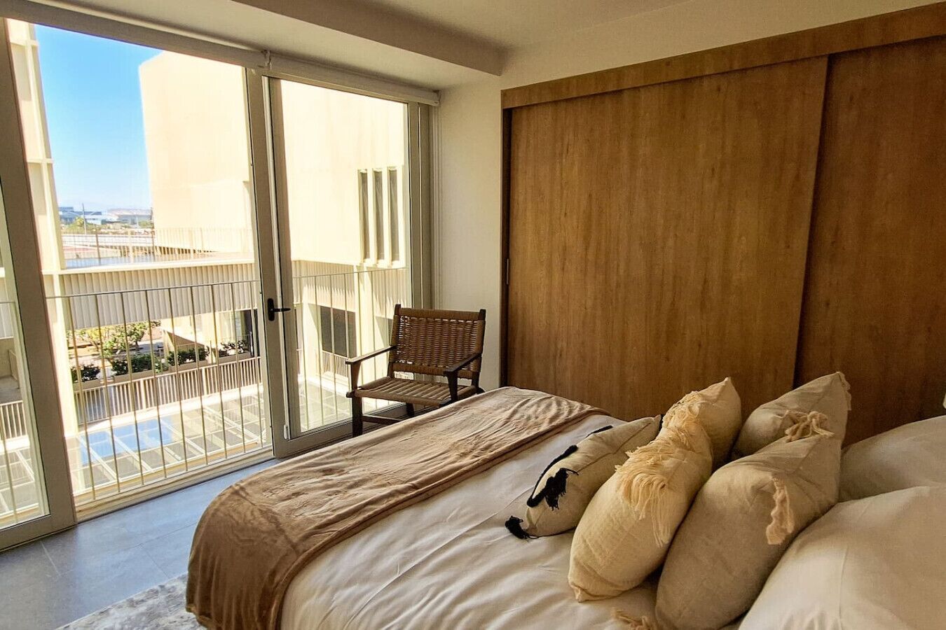Une chambre dotée d’un grand lit, d’une tête de lit en bois, d’une chaise et d’un balcon offrant une vue sur les bâtiments et la verdure.