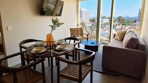 A modern living room with a dining area, a flat-screen TV mounted on the wall, and a large window offering a city view.