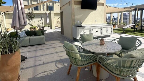An outdoor patio area with a round table, cushioned chairs, a large potted plant, and a TV mounted on a wall.