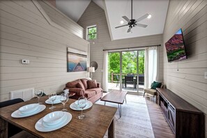 A dining area with a wooden table set for four, a brown sofa, and a television mounted on the wall. - Cozy Current Lake Cabin! +Pet Friendly +Game Loft! (Sevierville)