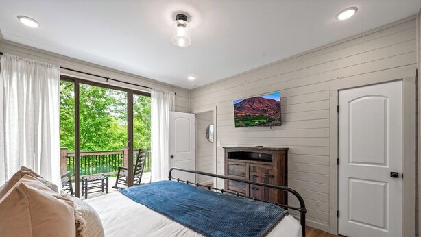A bedroom with a bed, a television, a fireplace, and a balcony with chairs. - Cozy Current Lake Cabin! +Pet Friendly +Game Loft! (Sevierville)