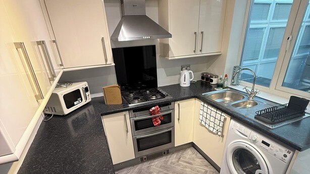 A modern kitchen with white cabinets, a black countertop, a stainless steel range hood, a microwave, a stove, a coffee maker, a sink, and a built-in washing machine. - 3 bedroom spacious home Parking and WiFi sleeps 5 (Bexleyheath)