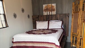 Une chambre à coucher avec un lit, une tête de lit en bois, un tableau, une fenêtre, une table de chevet, un vase avec des fleurs et une chaise en bambou.