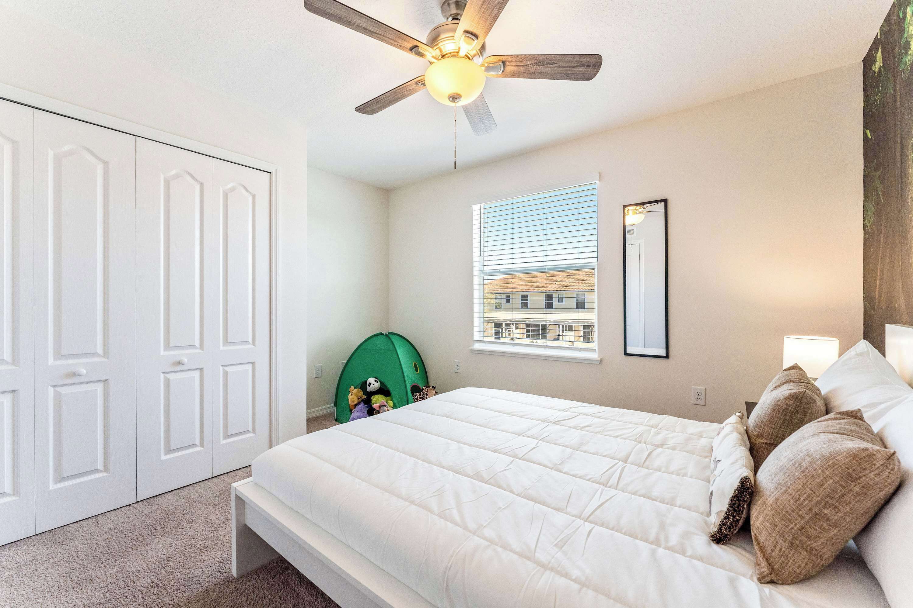 A bedroom with a bed, a ceiling fan, a window with blinds, a green play tent, and a white wardrobe.