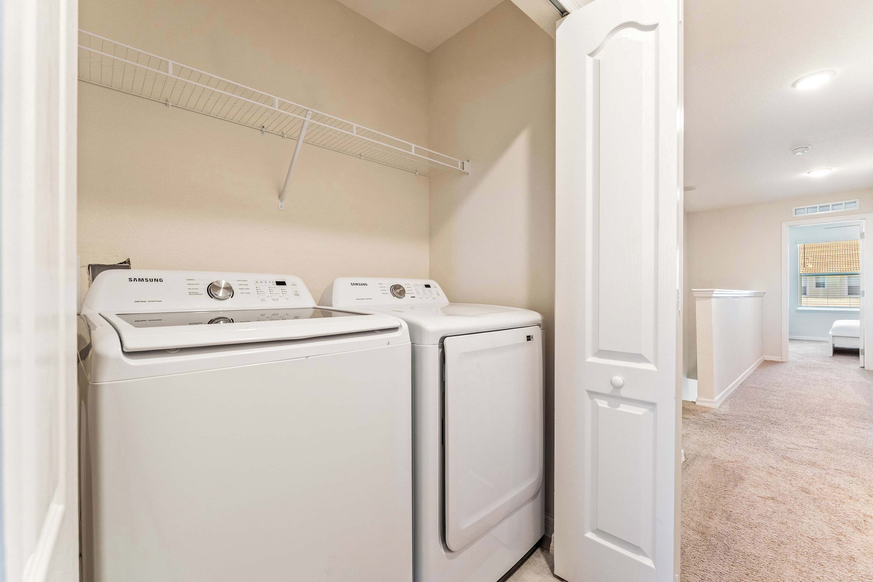 A laundry room with two white Samsung washing machines, a drying rack, and a door leading to another room.
