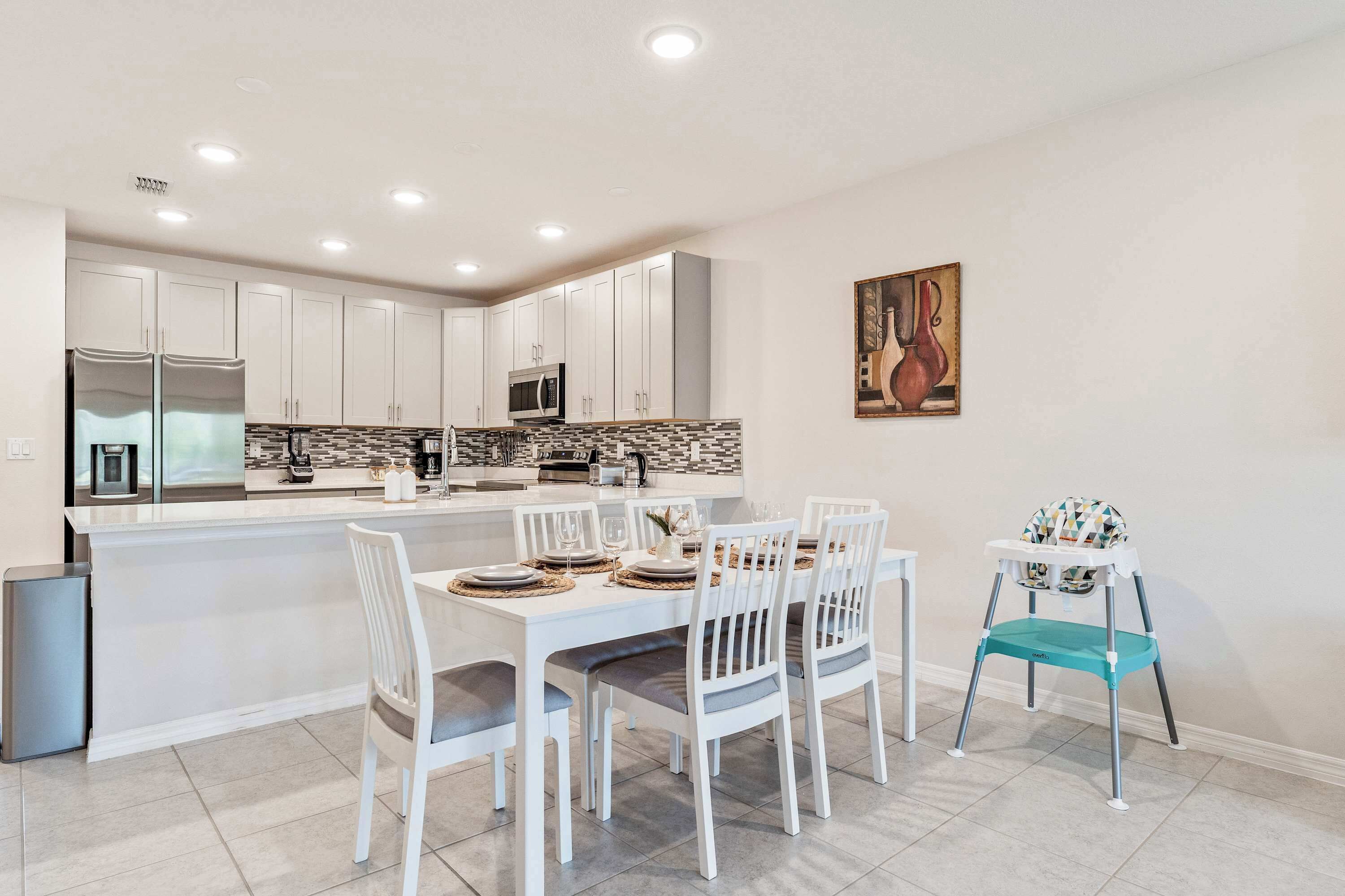 Une cuisine moderne avec des meubles blancs, un îlot central et un coin repas comprenant des chaises blanches et une chaise haute turquoise.