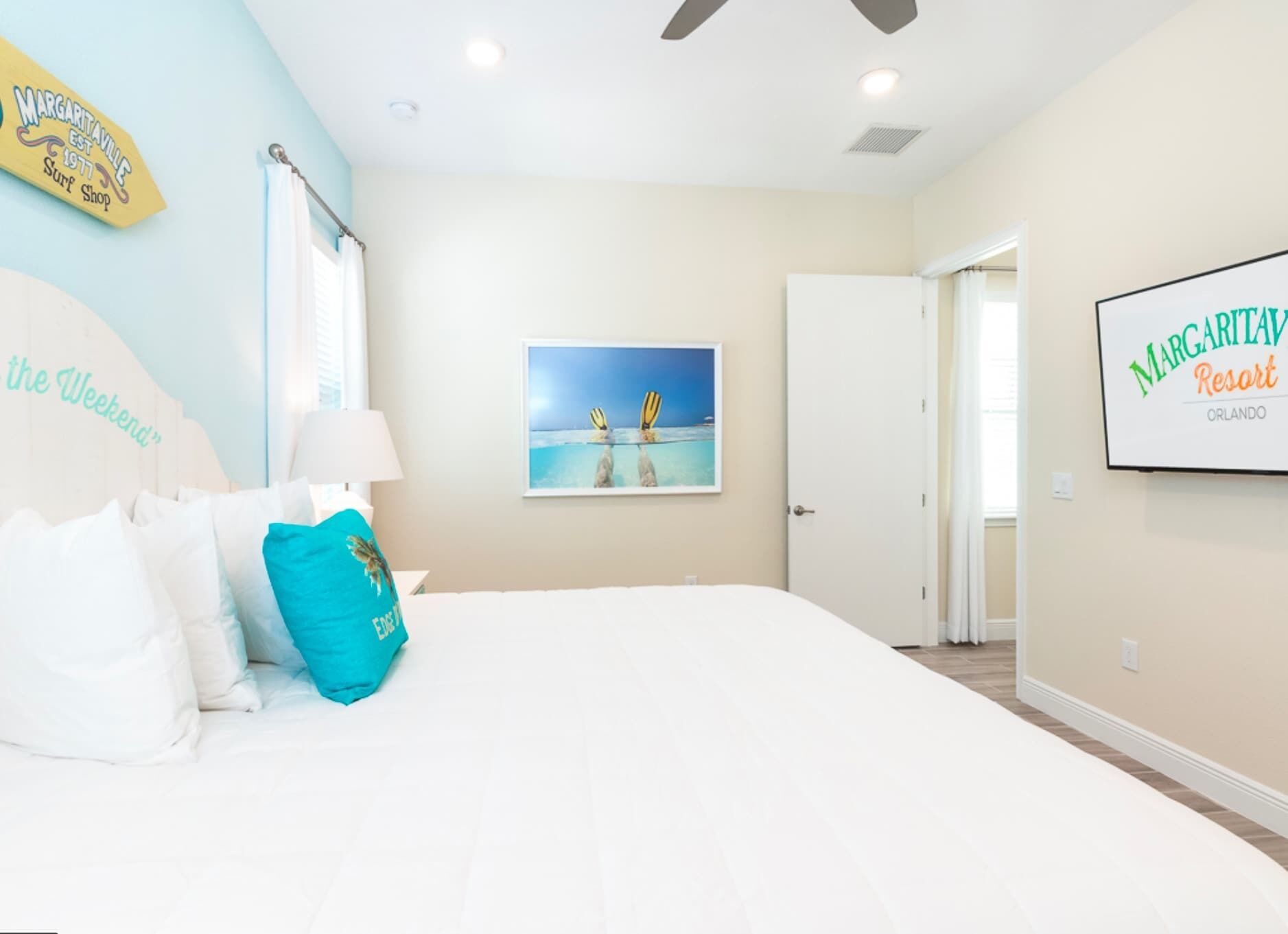 A bedroom with a bed, a ceiling fan, a painting of a beach scene, and a sign for Margaritaville Resort Orlando.