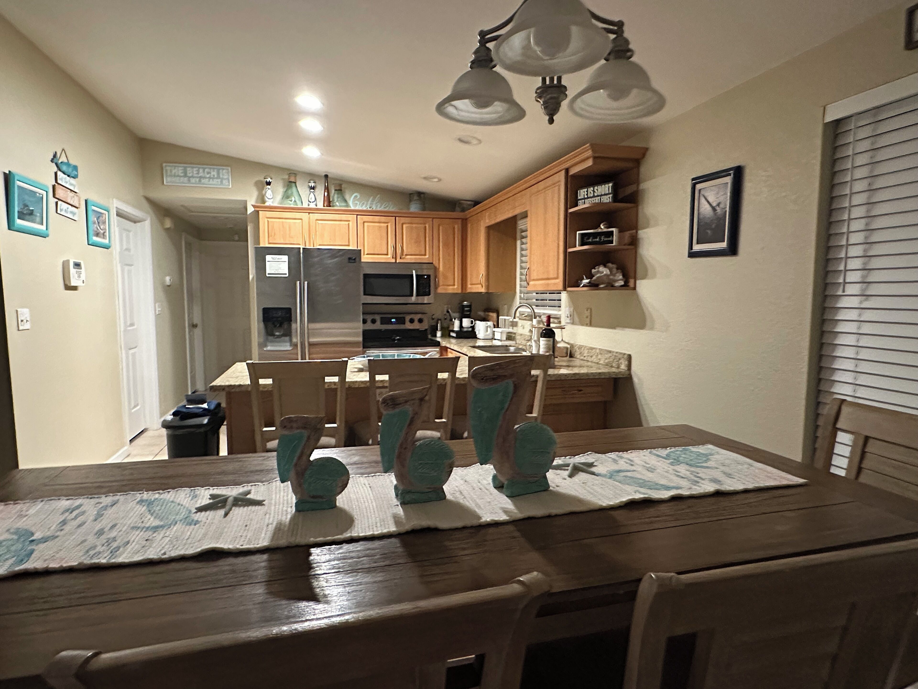 A kitchen with wooden cabinets, a stainless steel refrigerator, a microwave, and a sink. There is a dining table with chairs and a placemat.