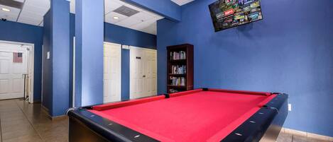 Une table de billard recouverte d’un drap de feutre rouge, une bibliothèque et un téléviseur fixé au mur diffusant les programmes d’ESPN.