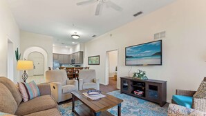 A living room with a sofa, armchair, coffee table, and a TV. There is a kitchen in the background.