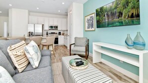 A modern living room with a grey sofa, a coffee table, and a TV on a stand. There is a kitchen in the background with white cabinets and a microwave. - Pistachio Palace Sleeps 10! (Kissimmee)