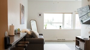 A spacious living room with a grey sofa, a coffee table, and a TV on a wooden stand. There are four bar stools and a ceiling fan. - Salted Roots Basecamp: 1 Bedroom Flat Downtown (Anchorage)