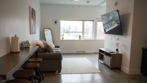 A modern living room with a sofa, bar stools, a flat-screen TV, and a large window. - Salted Roots Basecamp: 1 Bedroom Flat Downtown (Anchorage)