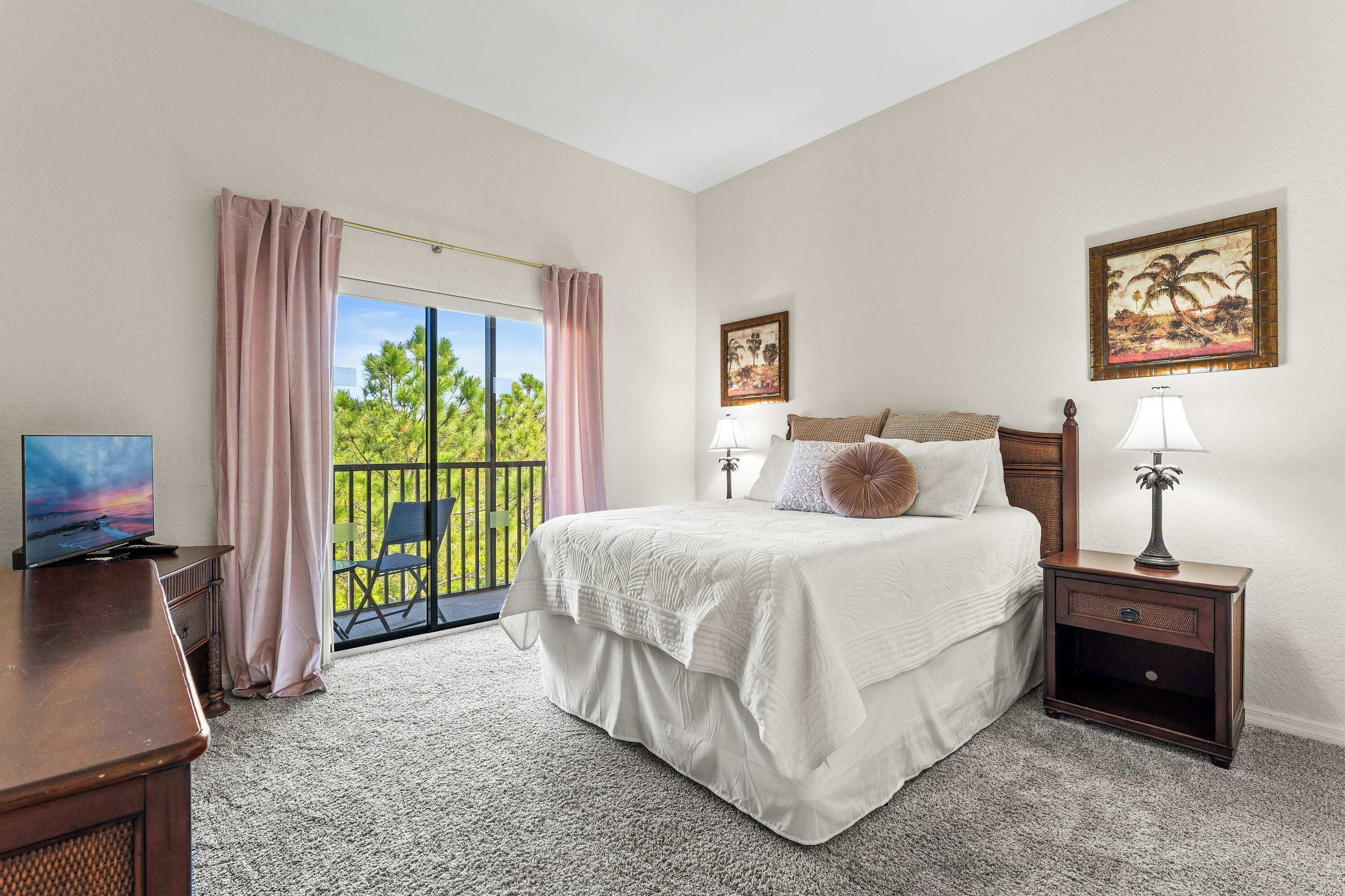 A bedroom with a bed, a nightstand, a desk, a TV, and a balcony with a view of greenery.