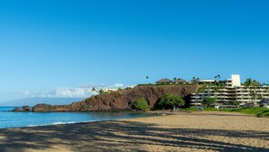 A beach with a rocky cliff, a clear blue sky, and a resort building with multiple floors and balconies. - NEW LISTING: Ka’anapali Royal K303 2BR – Ocean Views, Pool, Walk to Golf & Beach (Lahaina)
