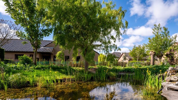 Un étang paisible aux eaux limpides, entouré d’une végétation luxuriante et de bâtiments traditionnels aux toits de tuiles.