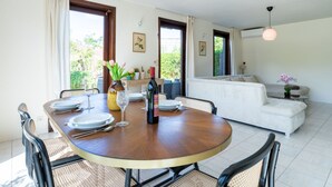 Un coin repas avec une table ronde en bois, des chaises et un vase de fleurs. On aperçoit un canapé et une petite table avec une lampe au fond.