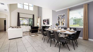 Una sala de estar moderna con un sofá blanco, sillas negras y una mesa de comedor preparada para seis comensales. Un televisor está montado en la pared, y hay un ventanal con cortinas.