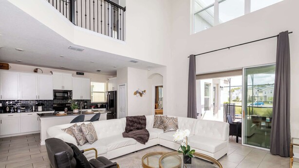 Un salón amplio con un sofá blanco, sillas negras y una mesa de centro de vidrio. Se ve una cocina en el fondo, con armarios blancos y electrodomésticos modernos.