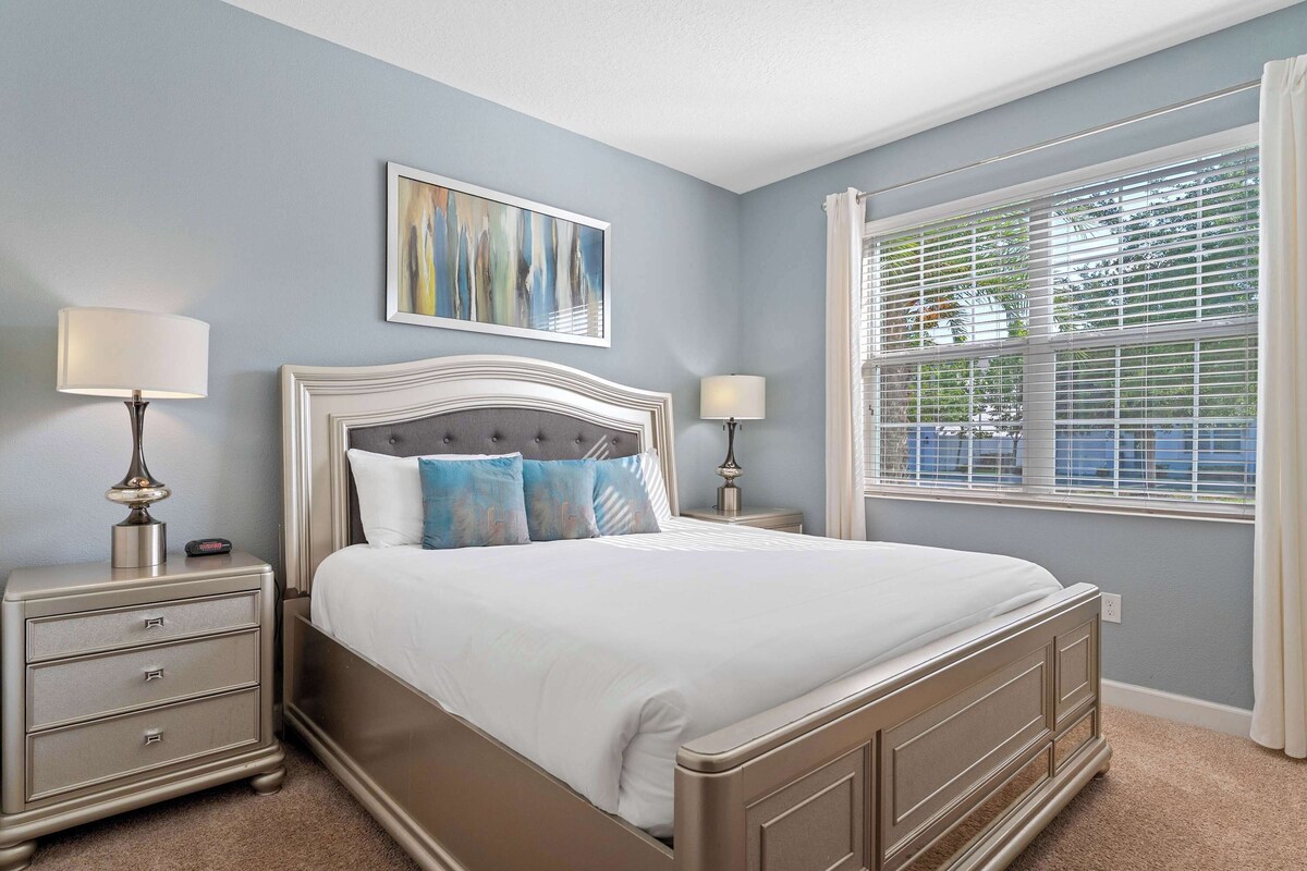 A bedroom with a bed, two bedside tables, a nightstand, a window with blinds, and a wall-mounted painting.