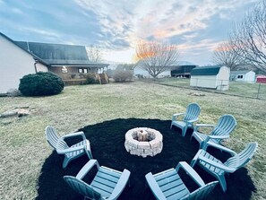 Un patio trasero con un hogar circular rodeado de sillas Adirondack, una terraza de madera y una casa al fondo.
