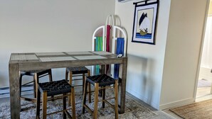 Une table à manger avec un plateau en bois et des pieds en métal, accompagnée de quatre tabourets noirs rembourrés. Un porte-planche de surf coloré est fixé au mur derrière la table.