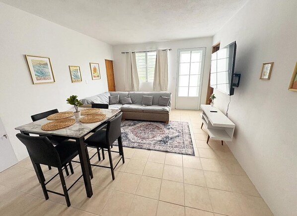 Un salon moderne comprenant un canapé, une table à manger avec des chaises, un téléviseur fixé au mur et une fenêtre avec des rideaux.
