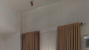Una habitación de hotel con cama, mesita de noche con lámpara y ventana con cortinas beige.